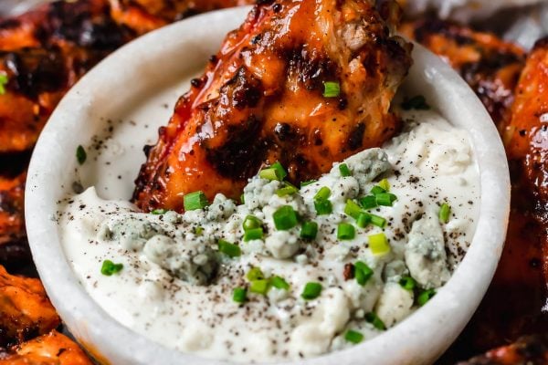 A crispy grilled wing dunked in a small bowl of bleu cheese dressing, in the center of a small baking sheet of grilled chicken wings, topped generously with snipped chives & served with bleu cheese dressing. The tray sits atop a black & white striped linen on a white surface next to a bottle of Franks RedHot hot sauce.