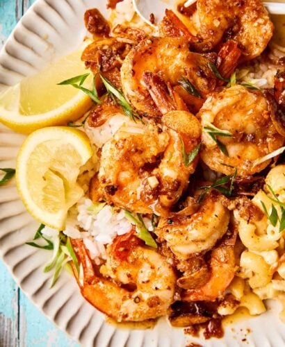 A plate of seasoned shrimp on white rice, garnished with green onions, served with lemon slices and macaroni and cheese on a white, ridged plate.