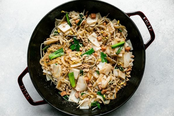 Top down photo of Hawaiian-style pork chow fun in a grenadine colored Staub cast iron wok. The wok sits on a light blue surface.