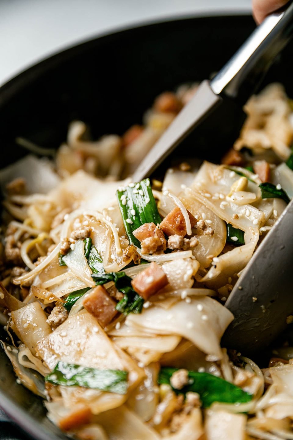 A close up of pork chow fun garnished with green onions and sesame seeds sitting inside of a wok. A pair of silicone tongs are picking up a scoop of the noodle stir fry.