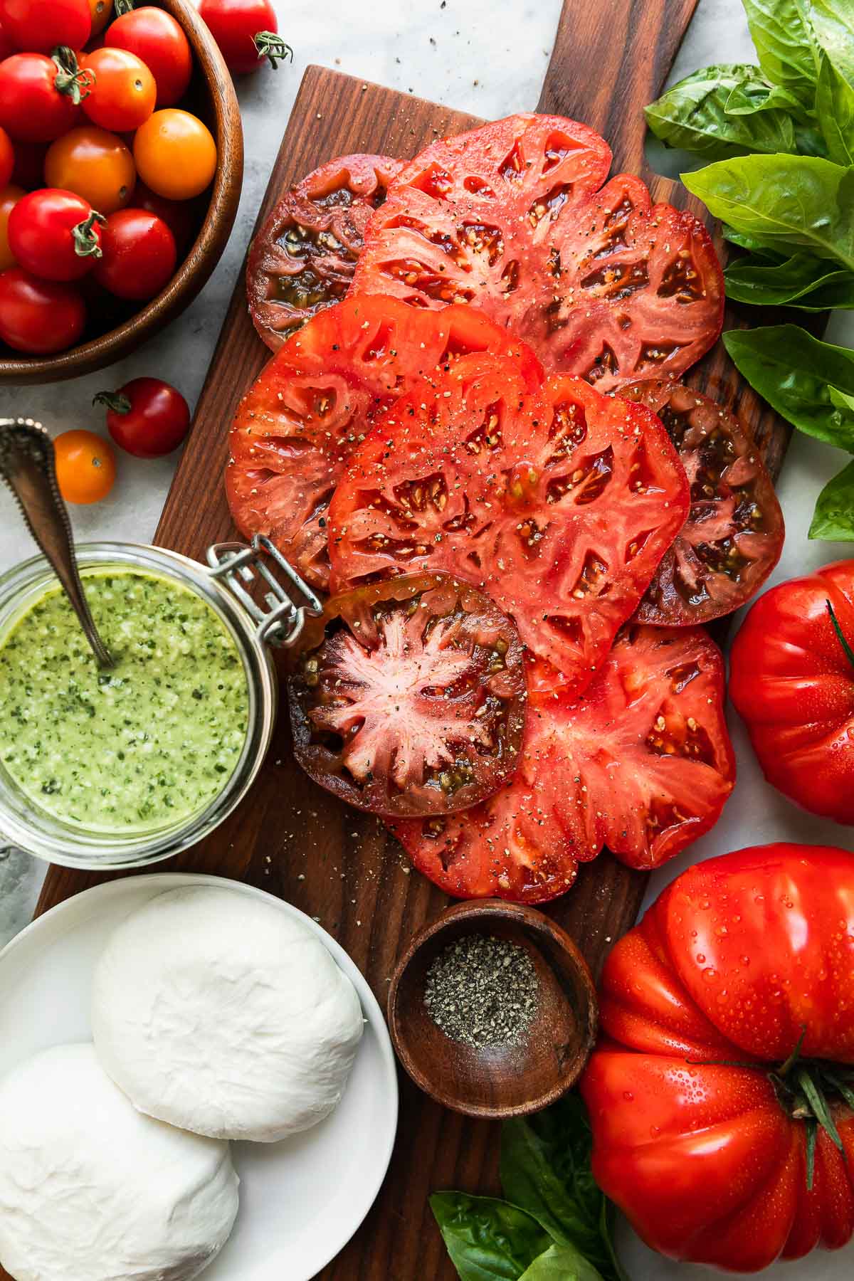 Tomato and burrata salad ingredients arranged atop a small wooden cutting board, in a small wood, glass, and ceramic bowls are arranged atop a white and gray marble surface: Heirloom tomatoes, whole cherry tomatoes, burrata cheese, basil pesto, ground black pepper, fresh basil.