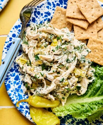 A blue speckled plate with creamy chicken salad mixed with herbs and chopped celery, served with lettuce, crackers, and a yellow pepper. A fork is on the side of the plate.