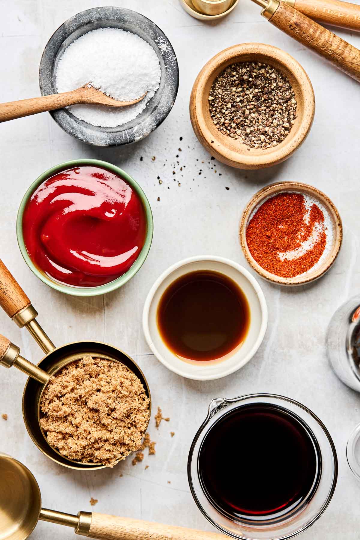 An overhead shot of ingredients in small bowls and measuring cups on a light grey textured surface: ketchup, brown sugar, shoyu, salt, pepper, cayenne pepper, and Worcestershire sauce.