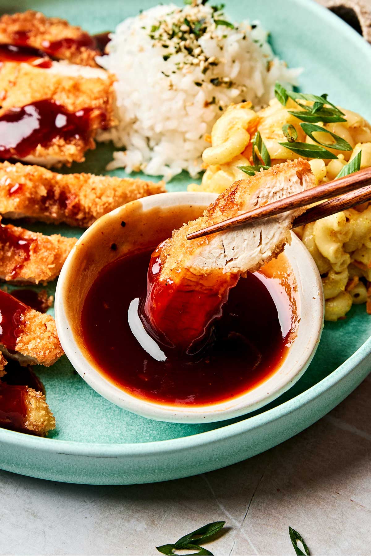 An angled shot of chopsticks dipping a piece of chicken katsu into a small white bowl of sauce on a light teal plate with katsu, white rice and mac salad.