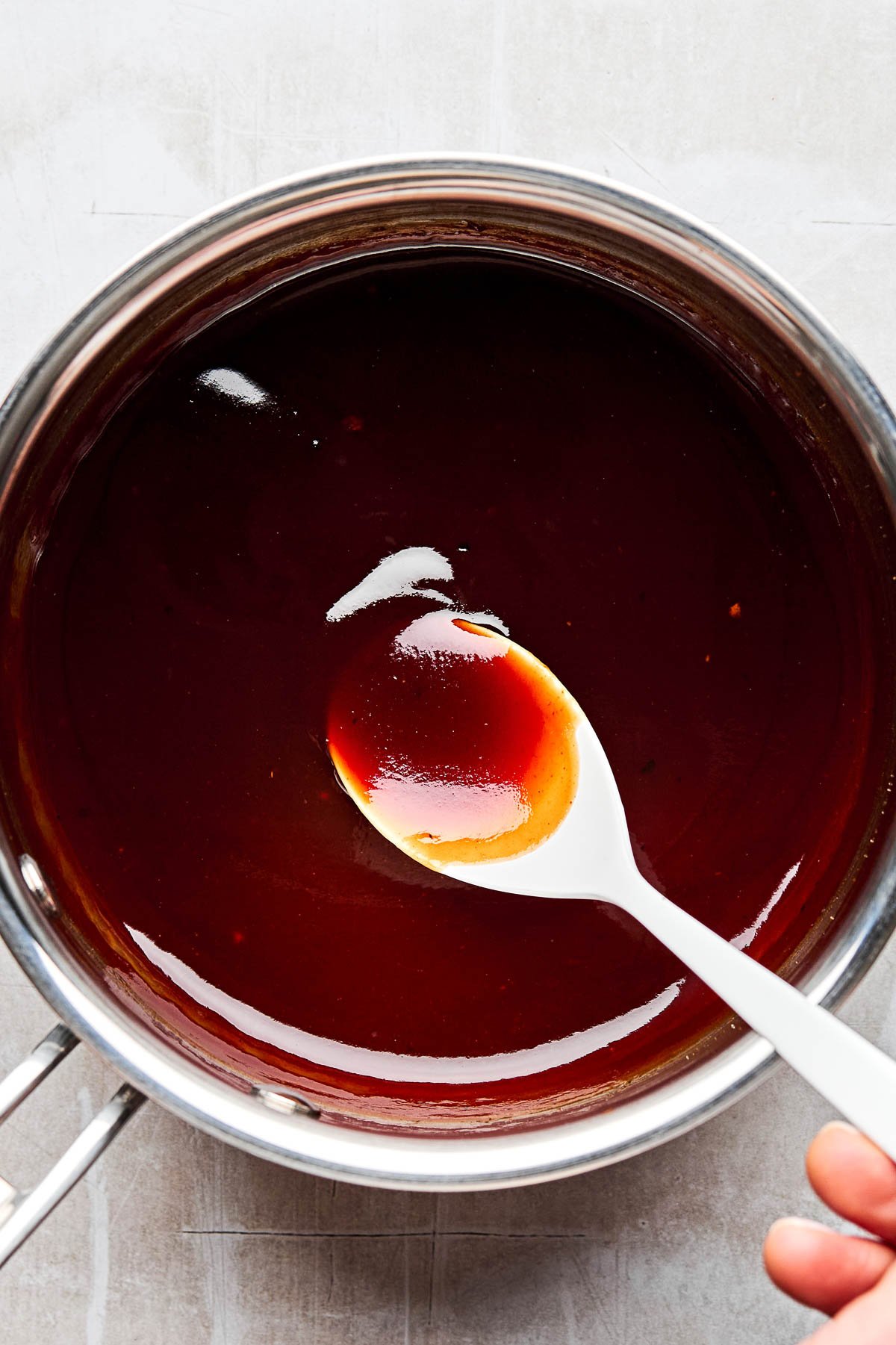 An overhead shot of a woman's hand holding up a spoonful of katsu sauce over a saucepan sitting atop a white textured surface.