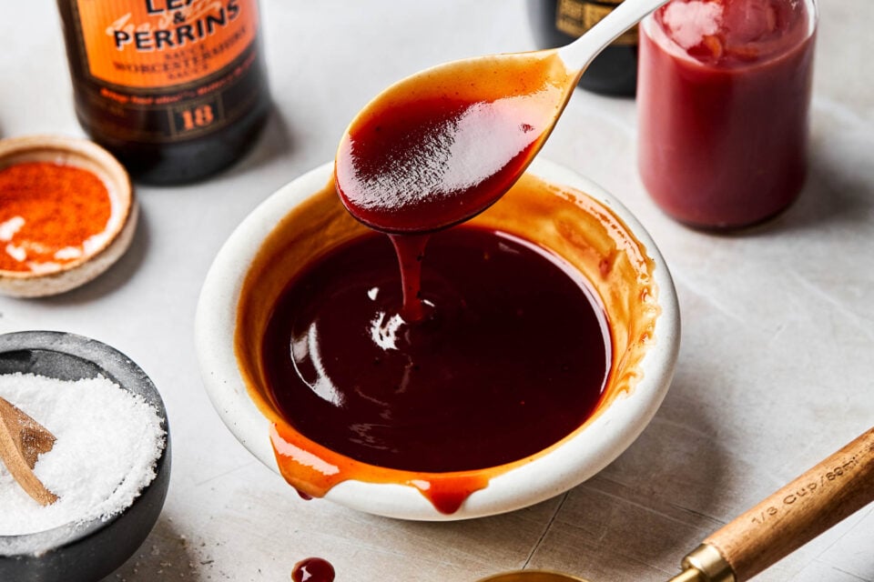 An angled shot of a spoon drizzling katsu sauce into a small bowl of sauce on a grey textured surface. The bowl is surrounded by containers of ketchup, brown sugar, shoyu, Worcestershire sauce, and sea salt.