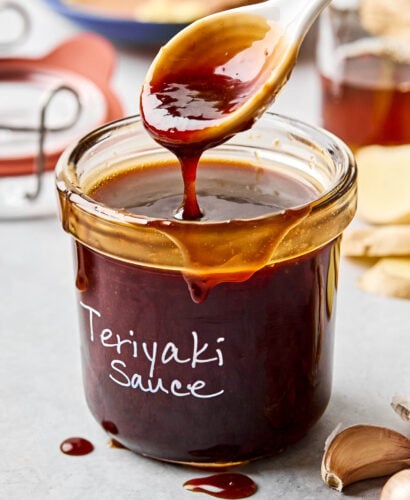 A side shot of a spoon drizzling teriyaki sauce into a labeled glass jar atop a white marbled surface. garlic, ginger, and bowls of ingredients can be seen in the background.