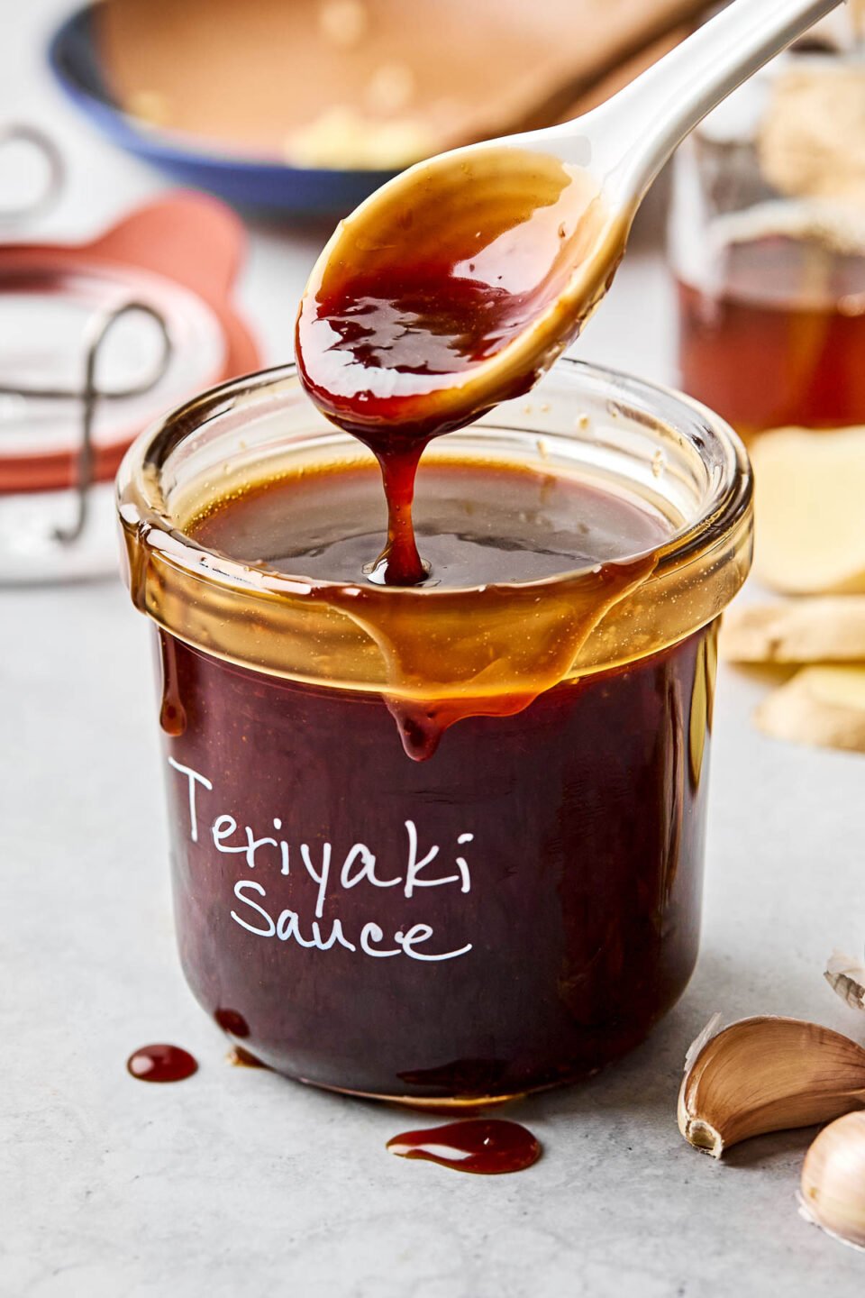 A side shot of a spoon drizzling teriyaki sauce into a labeled glass jar atop a white marbled surface. garlic, ginger, and bowls of ingredients can be seen in the background.