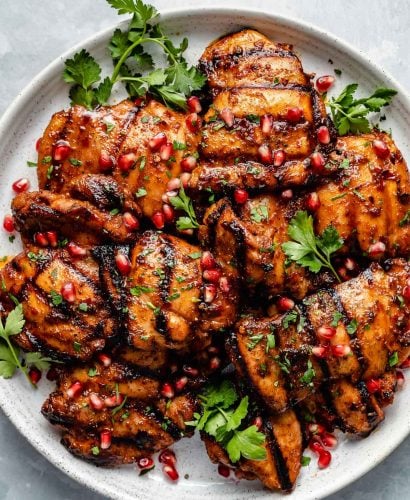 Grilled honey harissa marinated chicken thighs shown on a white speckled plate atop a light blue surface. The chicken is garnished with pomegranate arils & fresh cilantro leaves.