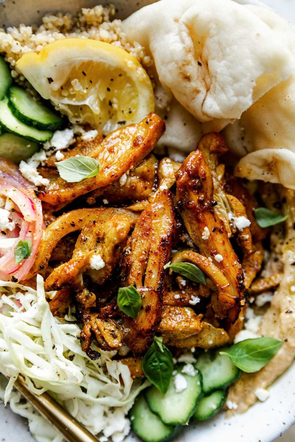Close up of chicken shawarma piled on top of a hummus bowl, along with shredded cabbage, sliced cucumber, pickled red onions, & naan bread.