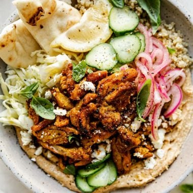 Chicken Shawarma Hummus Bowl with quinoa, cucumbers, pickled red onion, shredded cabbage, & naan bread shown in a speckled gray ceramic bowl. The bowl sits atop a creamy cement surface, surrounded by lemon wedges, fresh basil leaves & a small bowl of pickled red onions.