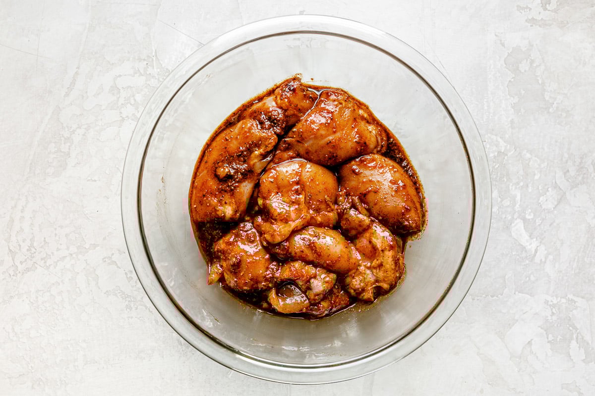 Chicken thighs marinating in shawarma marinade in a large clear mixing bowl atop a creamy white surface.