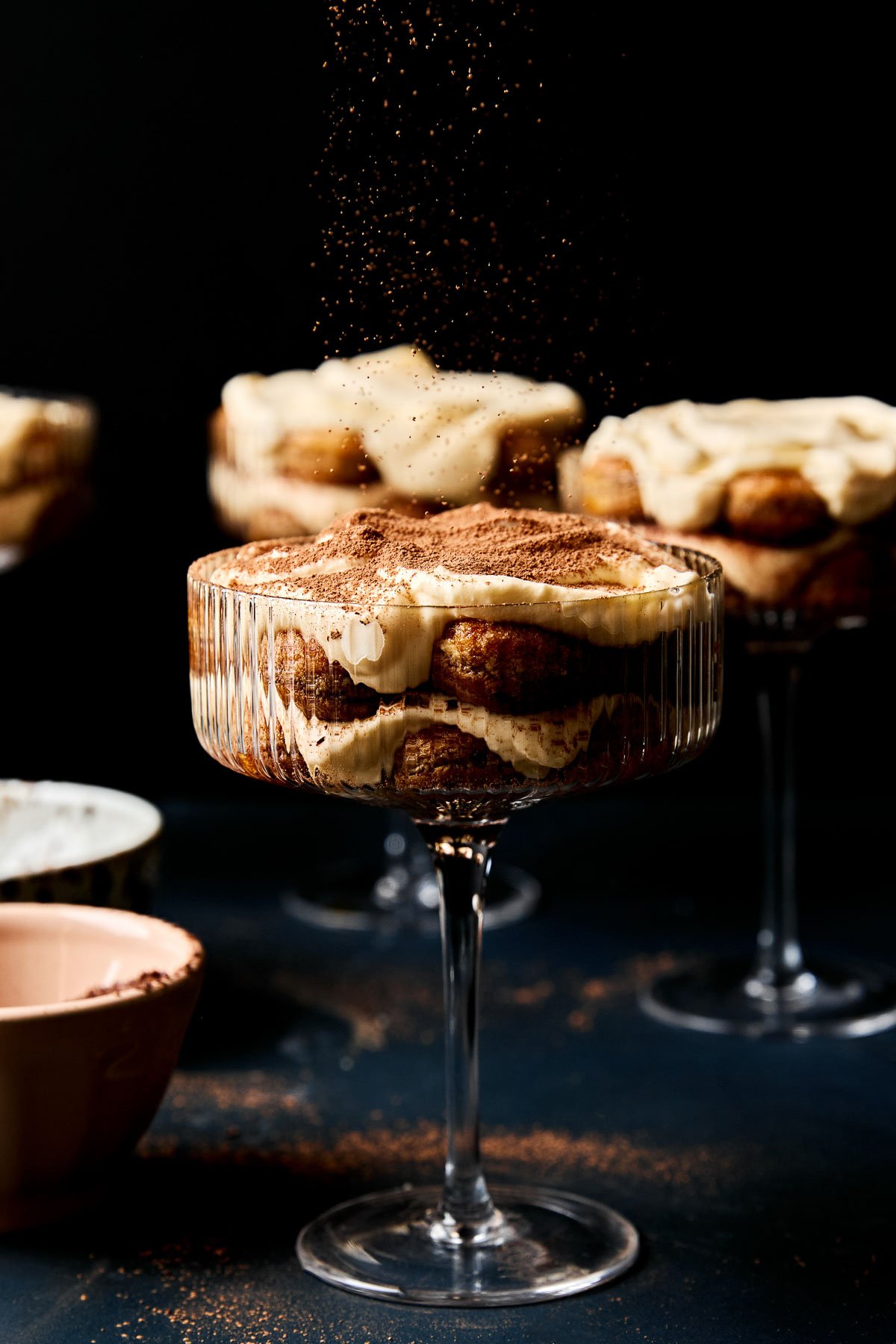 A side shot of cocoa powder being sprinkled over an individual tiramisu cup in a coupe glass. Three more tiramisu cups sit in the background on a black surface. 