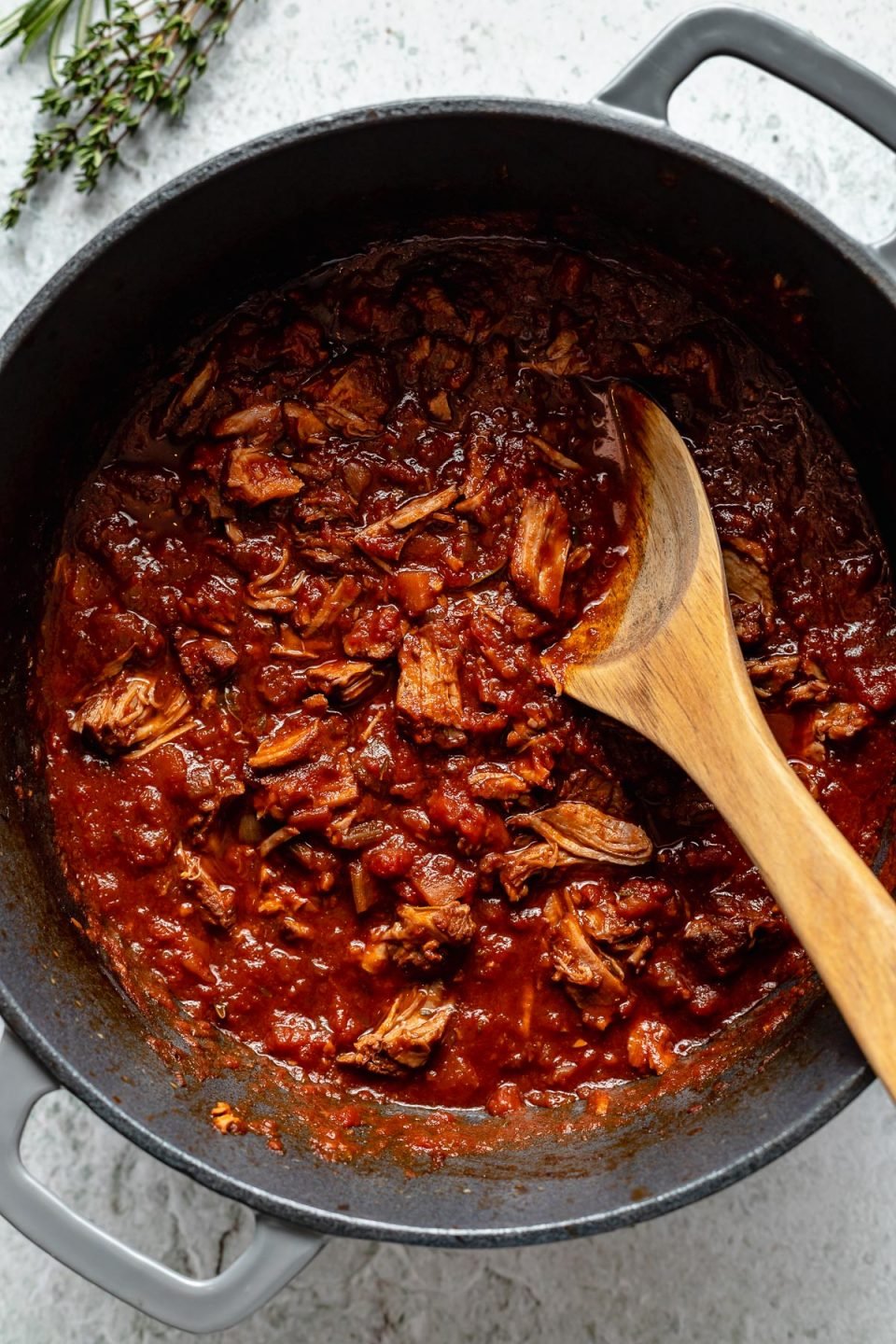 Braised lamb ragu in a large Dutch oven atop a light blue surface. There is a wooden spoon nestled in the ragu sauce & there are fresh herbs next to the pot.