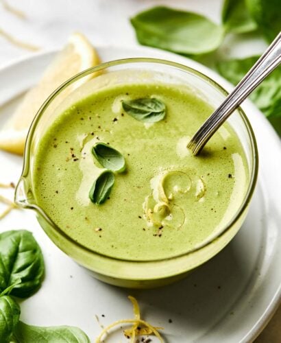 A glass bowl filled with creamy green dressing, garnished with fresh basil leaves, lemon zest, and black pepper. A metal spoon rests in the bowl, and basil leaves and a lemon wedge are on the white plate underneath.
