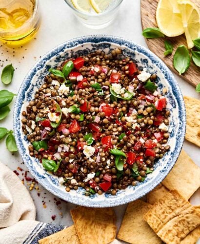 A blue and white bowl filled with lentil salad containing diced tomatoes, feta cheese, red onions, and fresh basil, surrounded by pita chips, lemon slices, and a glass of water with lemon.