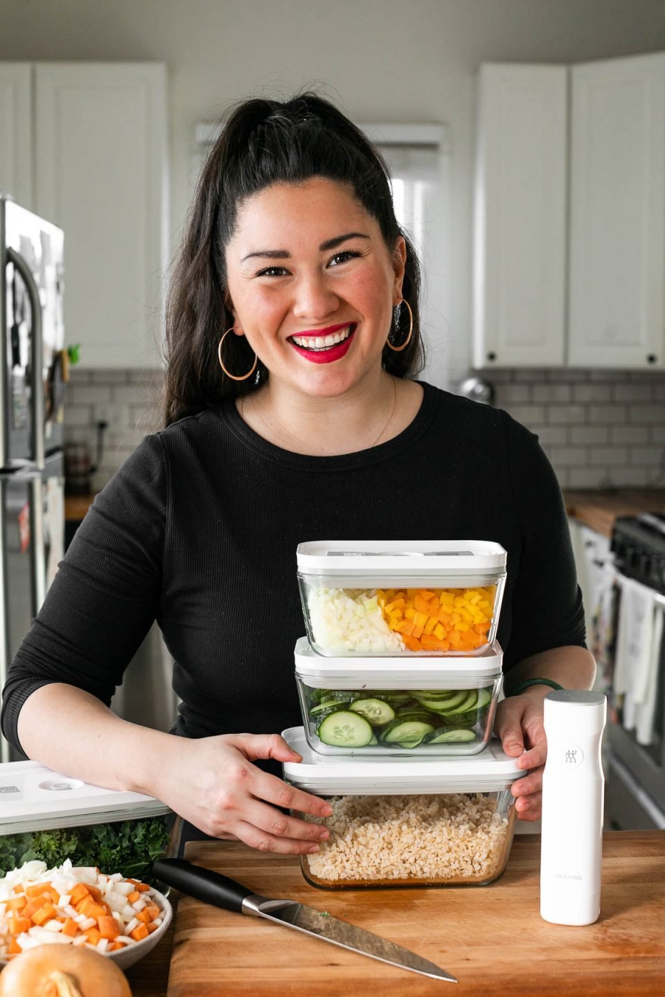 Jess standing at her kitchen counter holding 3 Zwilling Fresh & Save containers filled with rice, sliced cucumbers, onions & peppers. The containers sit atop a wood cutting board, next to a knife & some bowls filled with chopped veggies.