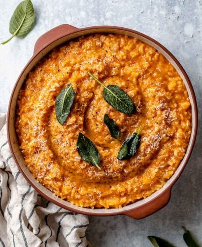 Creamy pumpkin risotto shown in a large terra cotta serving dish. The risotto is topped with crispy fried sage leaves & finely grated parmesan. The dish sits atop a light blue surface, next to a striped linen napkin & some fresh sage leaves.