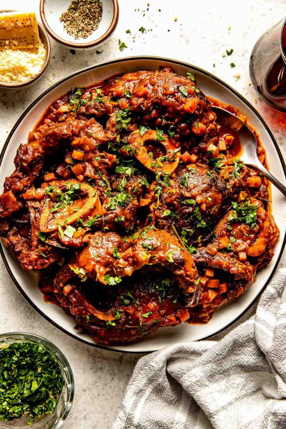 An overhead shot of prepared osso buco garnished with parsley on a white plate atop an off-white textured surface. The plate is surrounded by dishes of parsley, parmesan and pepper, and a glass of red wine.