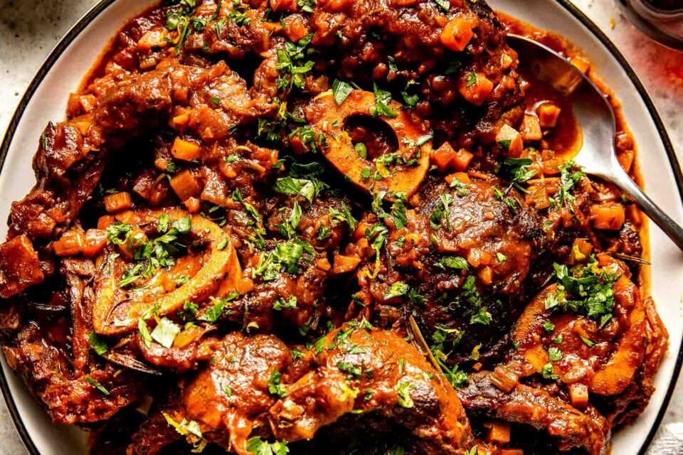 An overhead close-up shot of osso buco garnished with parsley on a white plate atop an off-white textured surface.
