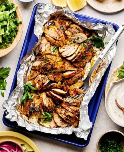 An overhead shot of sliced shawarma chicken thighs on a foil-lined blue sheet pan atop a white surface. The pan is surrounded by dishes of lemon wedges, diced tomatoes and cucumbers, shredded lettuce, pickled red onions, turmeric rice, shawarma sauce and fresh herbs.