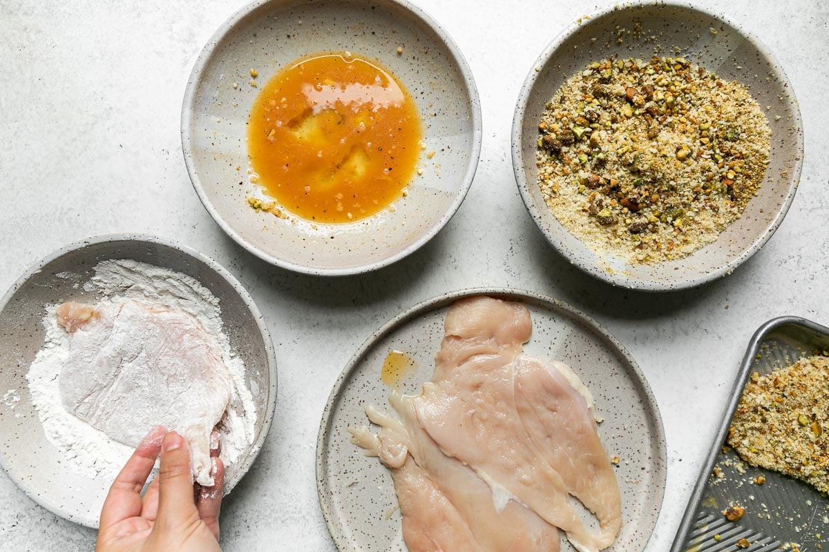 How to make Parmesan Pistachio Crusted Chicken, step 3: Bread the chicken. A woman's hand works to dip a single chicken breast into a gray ceramic bowl filled with all-purpose flour as she begins to work her way through a dredging station for pistachio crusted chicken. To complete the dredging station there is another bowl filled with whisked eggs, then a mixture of chopped pistachios, panko breadcrumbs, and grated Parmigiano Reggiano that fills a third bowl, and a plate topped with boneless, skinless chicken breasts. All sit atop a creamy white textured surface. Resting alongside is an aluminum baking sheet with one completely breaded pistachio chicken cutlet ready to be cooked.