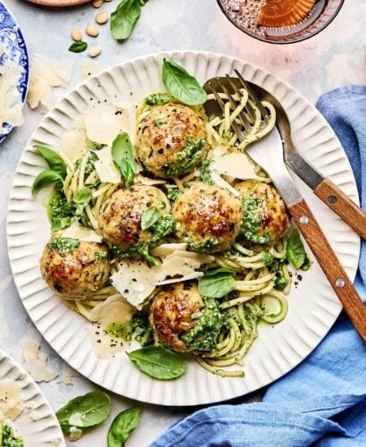 An overhead shot of turkey meatballs on a bed of pasta with pesto sauce on a white scalloped plate, garnished with basil leaves and shaved parmesan. The plate sits on a grey textured surface alongside a second dish of pasta, dishes of parmesan, pine nuts, basil leaves and tomatoes.