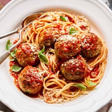 An angled shot of spaghetti and chicken meatballs in red sauce in a white shallow bowl atop a white marbled surface, surrounded by salt and pepper, a plate of bread and a glass of red wine.