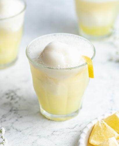 A close-up shot of a sgroppino cocktail atop a white marbled surface. A plate of lemon wedges and two additional cocktails sit beside it.