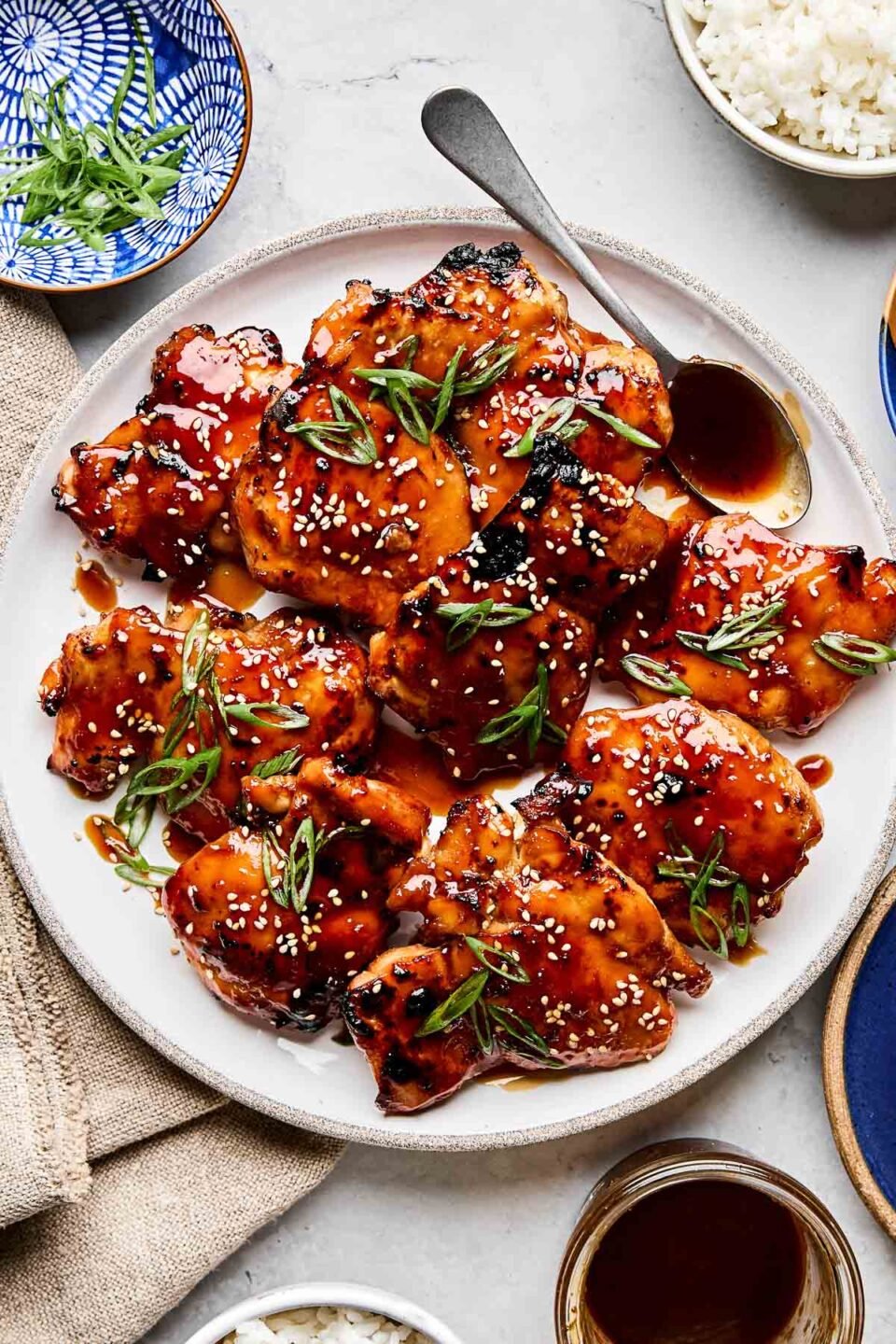 An overhead shot of teriyaki chicken garnished with sesame seeds and green onions on a white plate atop a white marbled surface. The plate is surrounded by dishes of rice, green onions, and teriyaki sauce.