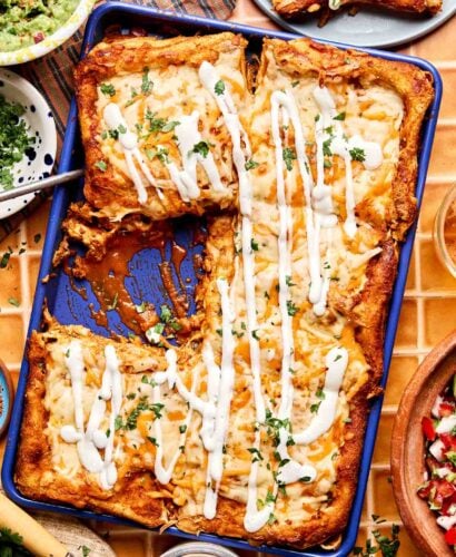 A blue baking dish filled with cheesy enchiladas topped with white sauce and chopped herbs, sitting on a tiled table surrounded by bowls of guacamole, pico de gallo, and avocado slices.