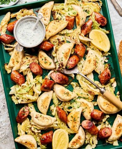 A green baking sheet with baked pierogis, sliced sausage, roasted cabbage, lemon wedges, and a small bowl of creamy dipping sauce garnished with chopped herbs. A serving fork and spoon rest on the tray.