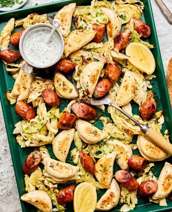 A green baking sheet with baked pierogis, sliced sausage, roasted cabbage, lemon wedges, and a small bowl of creamy dipping sauce garnished with chopped herbs. A serving fork and spoon rest on the tray.