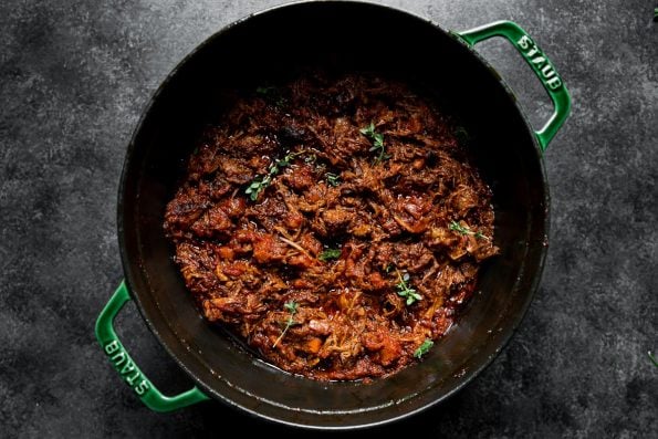 Slowly braised beef short rib ragu in a large Dutch oven atop a dark textured surface. A few fresh herbs garnish the top of the ragu sauce.