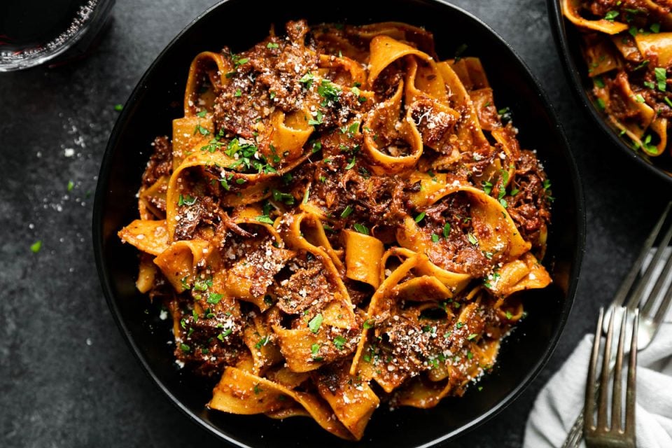 Braised Short Rib Ragu Pappardelle shown in pasta bowls, topped with grated parmesan & fresh parsley. The bowls sit atop a black surface surrounded by a clear glass filled with red wine, a gray linen napkin, & two silver forks.