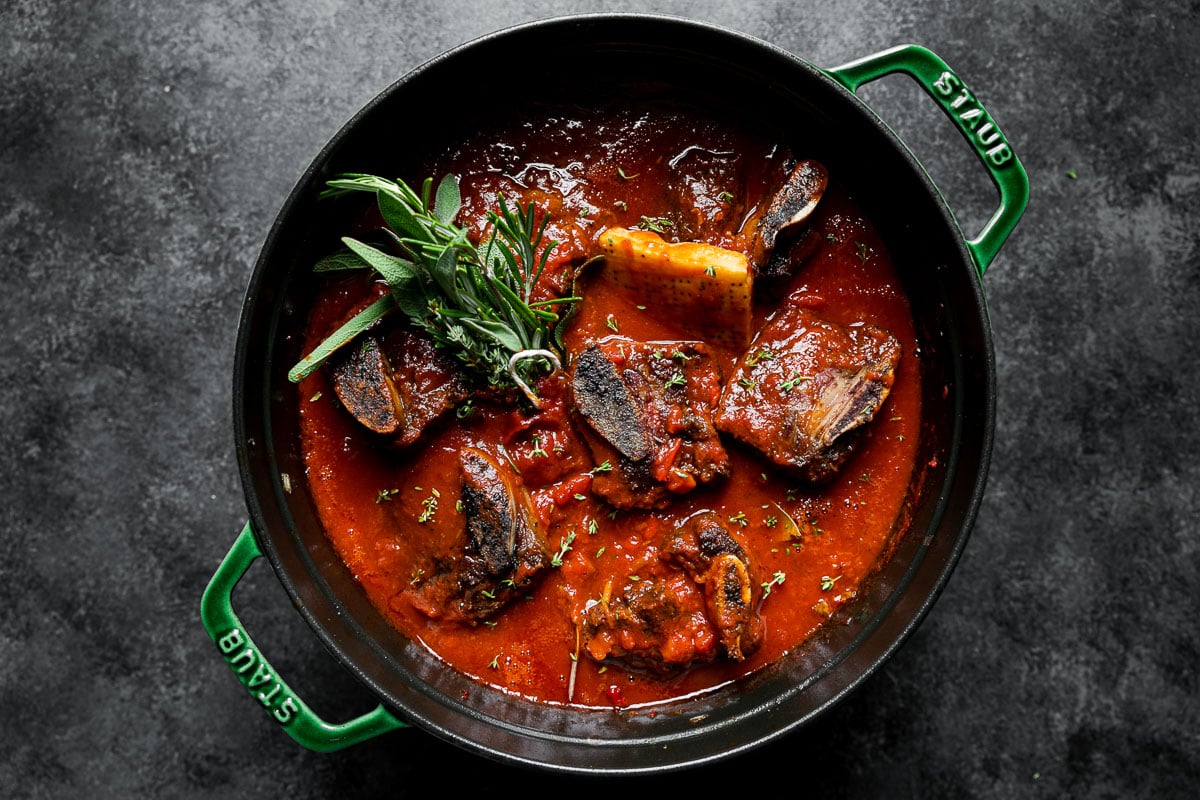 Beef short rib ragu sauce prior to braising - browned beef short ribs, fresh herbs, & a parmesan rind sitting in a crushed tomato mixture in a large Dutch oven atop a dark textured surface.