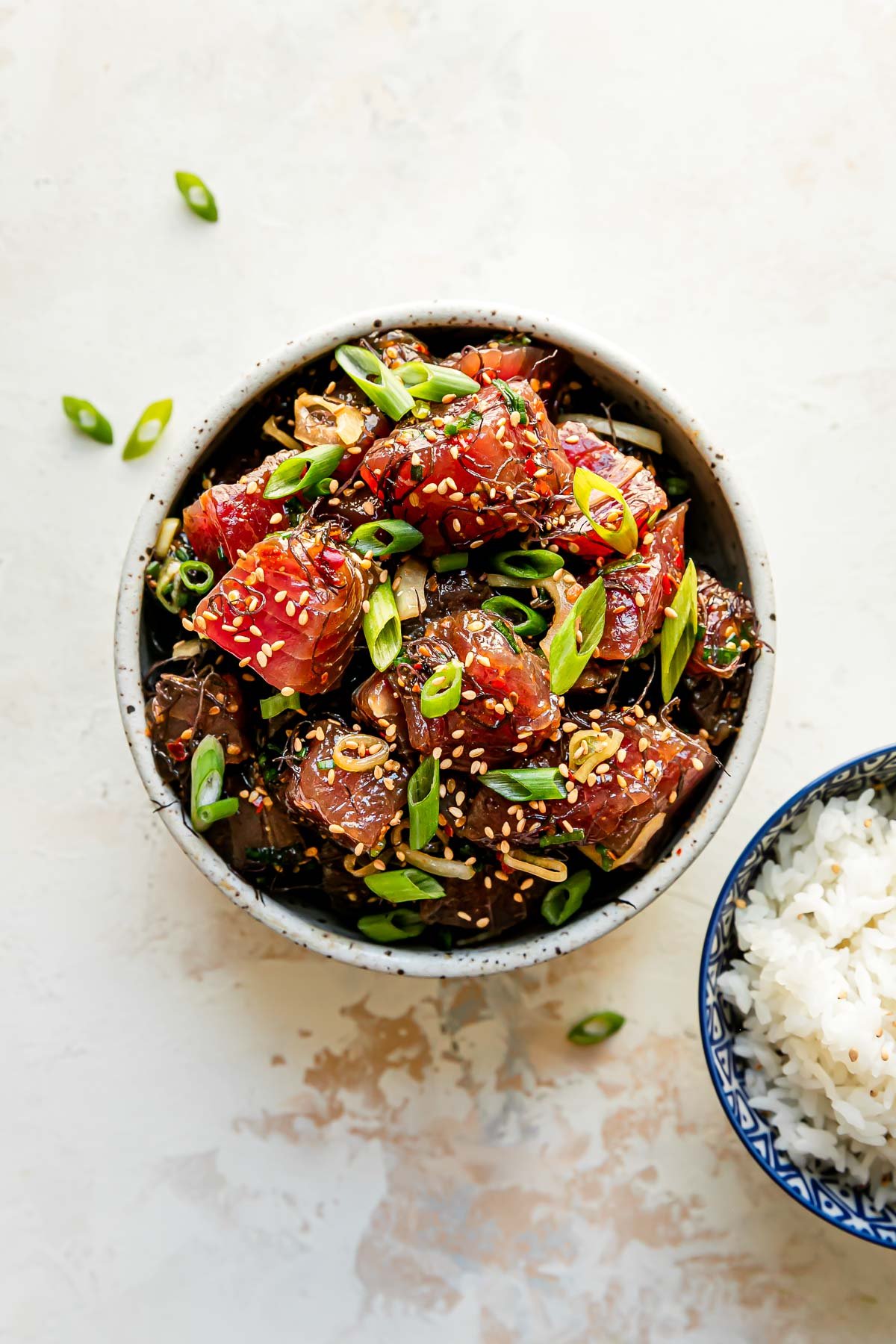 Shoyu poke fills a small ceramic bowl that sits atop a creamy white textured surface. It is garnished with toasted sesame seeds and thinly sliced green onion. A small blue and white bowl filled with white rice rests alongside the bowl of tuna poke at center.