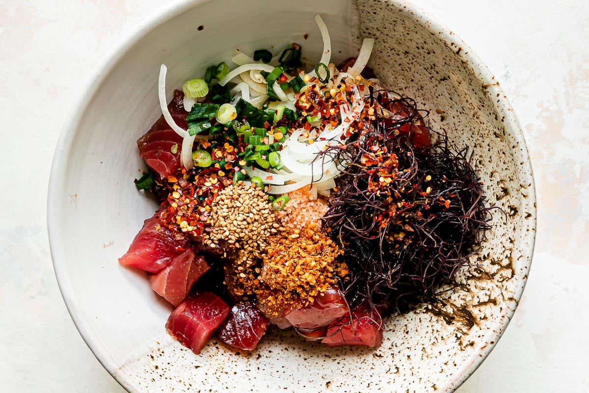 Tuna poke components are arranged in a large ceramic bowl that sits atop a creamy white textured surface: ahi tuna, sweet onion, green onions, ogo or limu, shoyu, toasted sesame oil, Hawaiian sea salt, Inamona, toasted sesame seeds, and crushed red pepper.