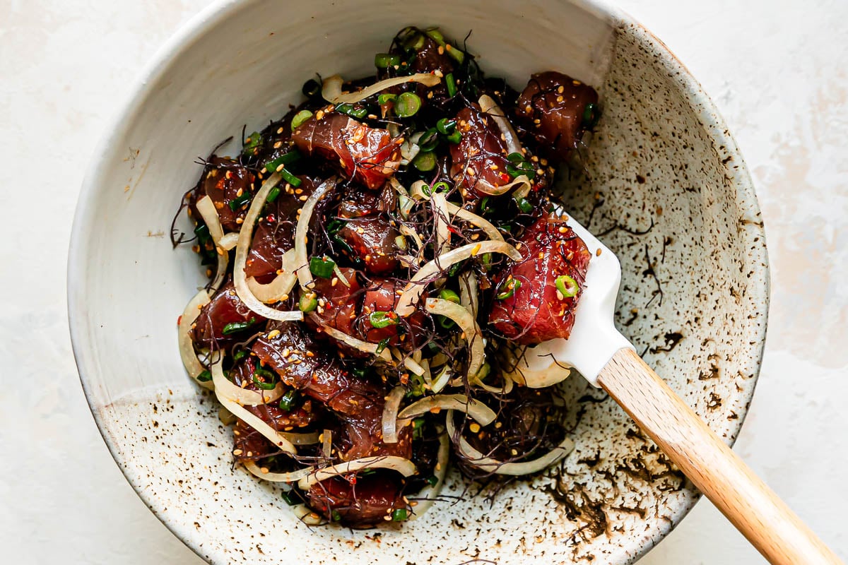 Shoyu ahi poke fills a large ceramic bowl that sits atop a creamy white textured surface. A spatula rests inside of the bowl for mixing.