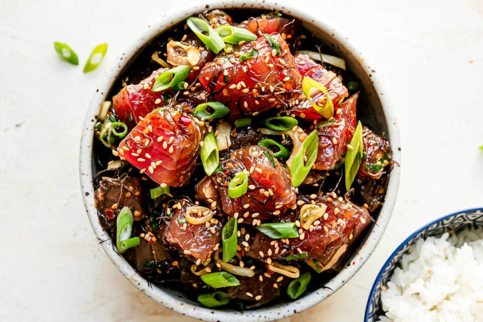 Shoyu poke fills a small ceramic bowl that sits atop a creamy white textured surface. The ahi poke has been garnished with toasted sesame seeds and thinly sliced green onion. A small blue and white bowl filled with white rice rests alongside the bowl of tuna poke at center.