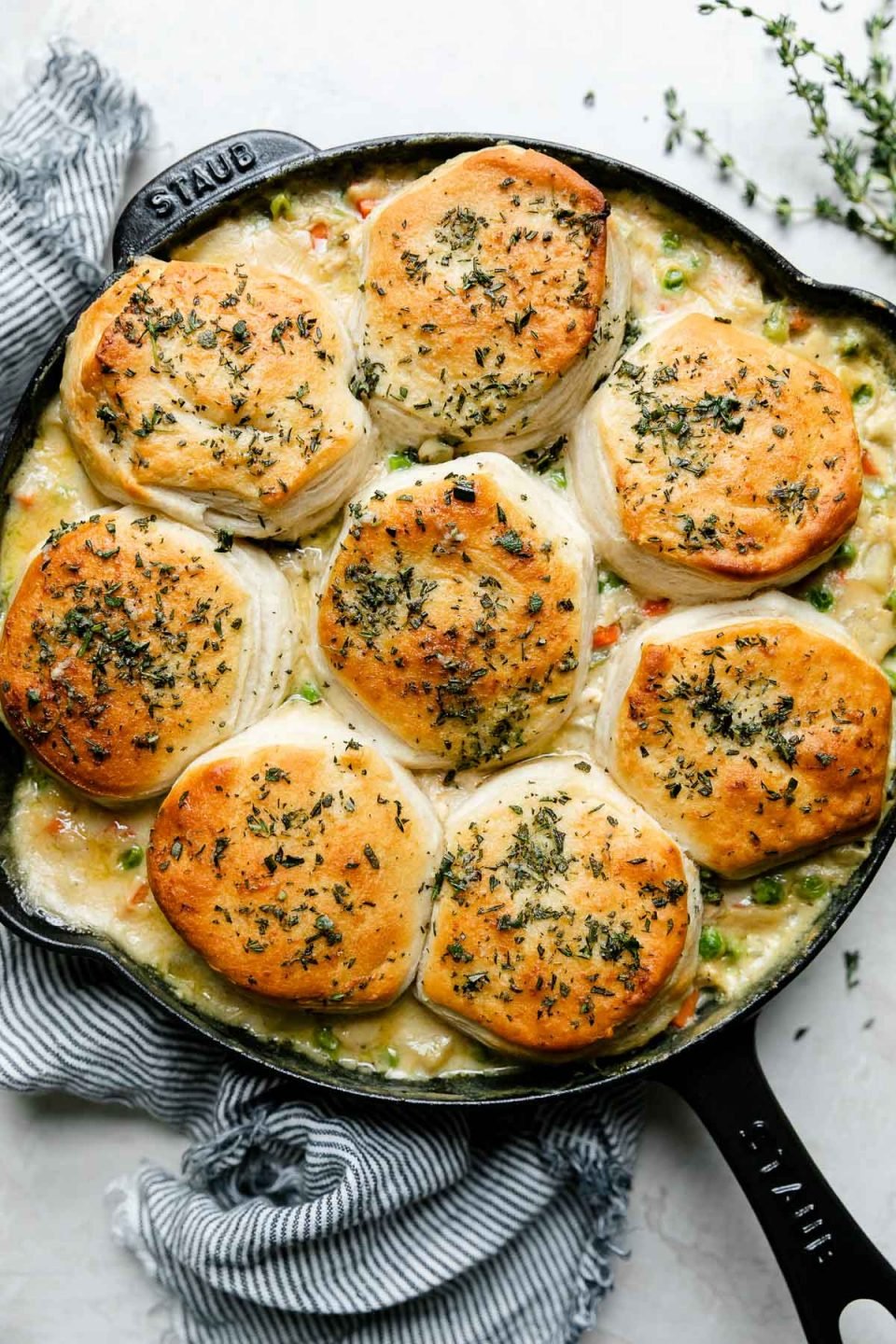 An overhead shot of skillet pot pie topped with biscuits in a black Staub cast iron skillet that sits atop a creamy white textured surface. A blue and white striped linen napkin rests alongside the skillet and sprigs of fresh thyme also surround the skillet.