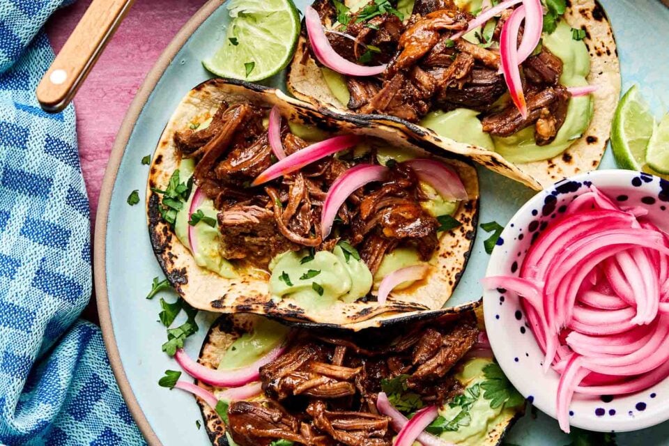 An overhead shot of a light blue plate holding three braised short rib tacos garnished with pickled red onions and fresh cilantro on a pink surface. A blue cloth sits alongside the plate.