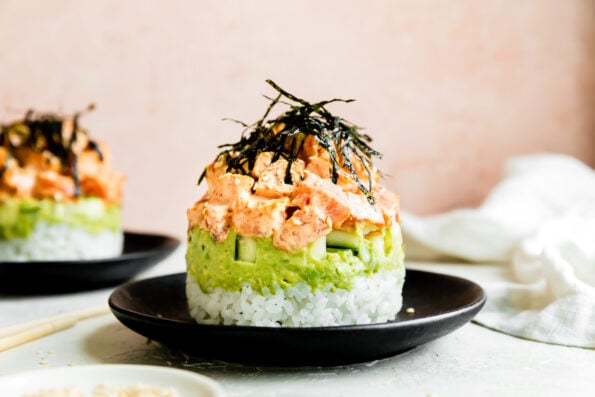 A sushi stack is served atop a black ceramic plate that sits atop a creamy white textured surface. The spicy tuna sushi stack has been garnished with a haystack of thinly sliced nori and a sprinkling of toasted sesame seeds. The plate is surrounded by a pair of chopsticks, a small white plate filled with toasted sesame seeds, a white linen napkin, and a second sushi stack served on another black plate rests in the background out of focus.