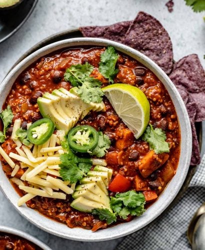 Sweet Potato Quinoa Chili shown in 2 soup bowls, topped with avocado, jalapeno, shredded vegan cheese, lime wedges & cilantro leaves. The bowls sit atop a light blue surface, next to a checkered blue & white linen, soup spoons, garnishes (sliced jalapeno, lime wedges, tortilla chips, etc.).