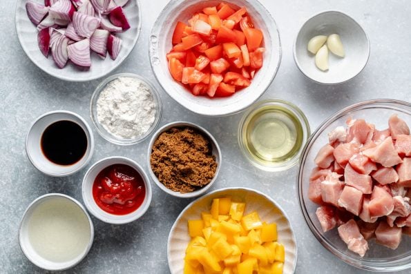 An overhead shot of ingredients arranged on a light blue surface in individual bowls and dishes: red onion, rice flour, soy sauce, ketchup, brown sugar, rice vinegar, oil, garlic, tomatoes, bell peppers, and diced pork loin.