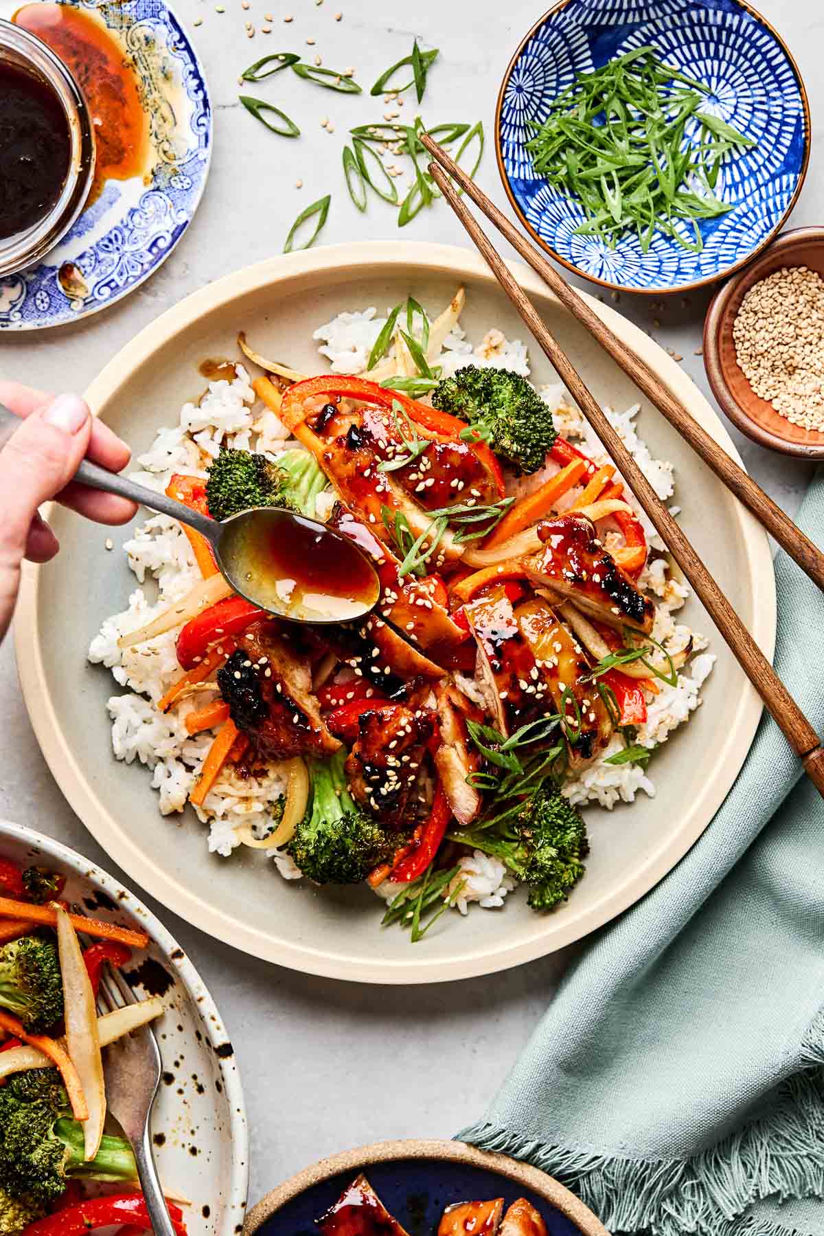 An overhead shot of teriyaki chicken and veggies served on a bed of rice and garnished with green onions and sesame seeds on an off-white plate with wooden chopsticks. A woman's hand drizzles teriyaki sauce from a spoon onto the bowl. The bowl is surrounded by dishes of green onions, sesame seeds, teriyaki sauce and an additional teriyaki chicken bowl.