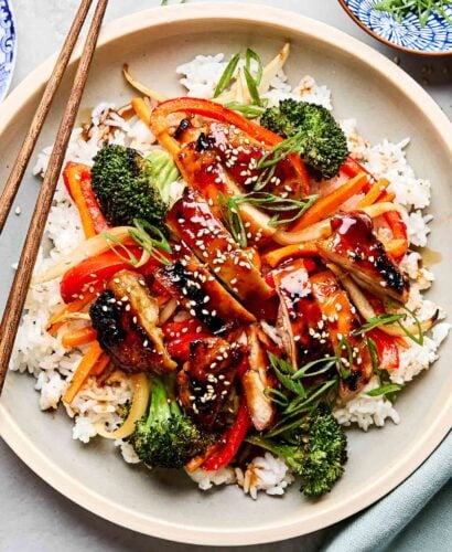 An overhead shot of teriyaki chicken and veggies served on a bed of rice and garnished with green onions and sesame seeds on an off-white plate with wooden chopsticks. The plate sits on a white marbled surface alongside a blue cloth and a small dish of green onions.