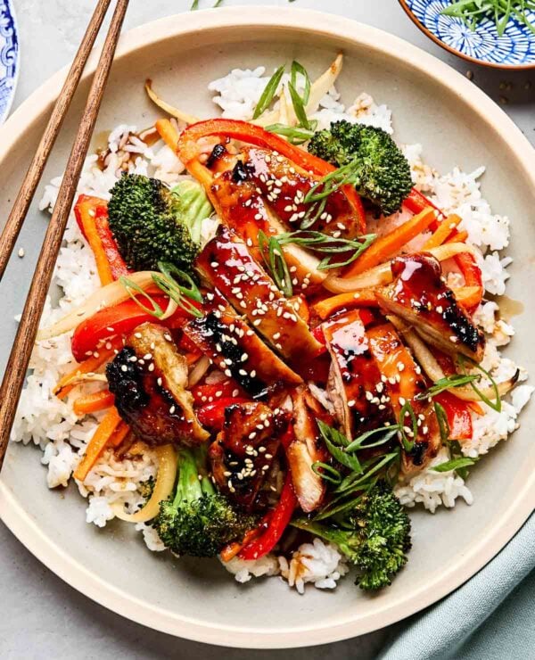 An overhead shot of teriyaki chicken and veggies served on a bed of rice and garnished with green onions and sesame seeds on an off-white plate with wooden chopsticks. The plate sits on a white marbled surface alongside a blue cloth and a small dish of green onions.