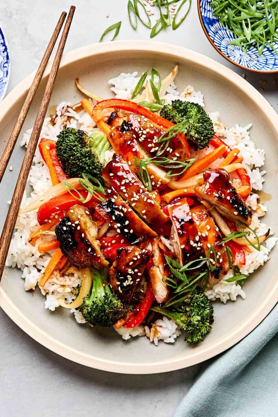 An overhead shot of teriyaki chicken and veggies served on a bed of rice and garnished with green onions and sesame seeds on an off-white plate with wooden chopsticks. The plate sits on a white marbled surface alongside a blue cloth and a small dish of green onions.