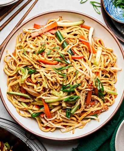 An overhead shot of stir fried vegetable noodles on a white stoneware plate on a white marbled surface. The plate is surrounded by a wok of noodles and a small blue dish of sliced green onions.